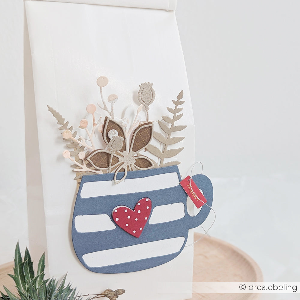Ein gestreifter Pappbecher mit einem Herz und getrockneten Blumen, gefertigt aus erlesenen Designpapieren, ziert eine weiße Geschenktüte auf einem Holztablett.