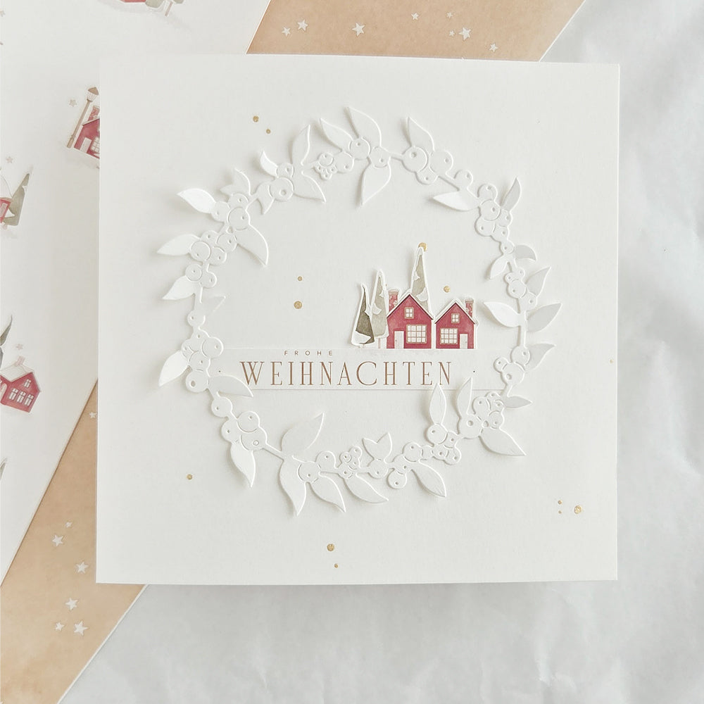 Eine quadratische Weihnachtskarte mit einem weißen „Motivstanze Beerenkranz“ und kleinen roten Häuschen sowie dem Schriftzug „Frohe Weihnachten“.