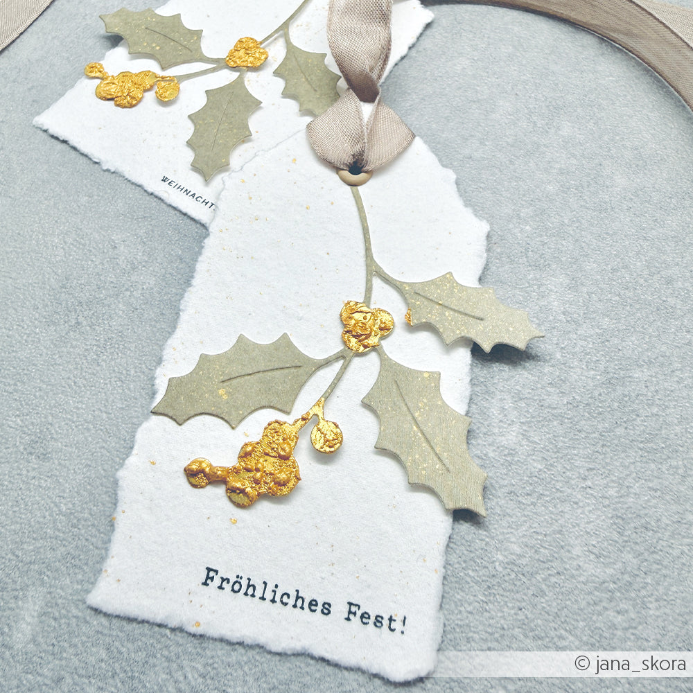 Zwei handgefertigte Geschenkanhänger aus Designpapier mit goldenen Stechpalmenzweigen und dem deutschen Text Fröhliches Fest!.