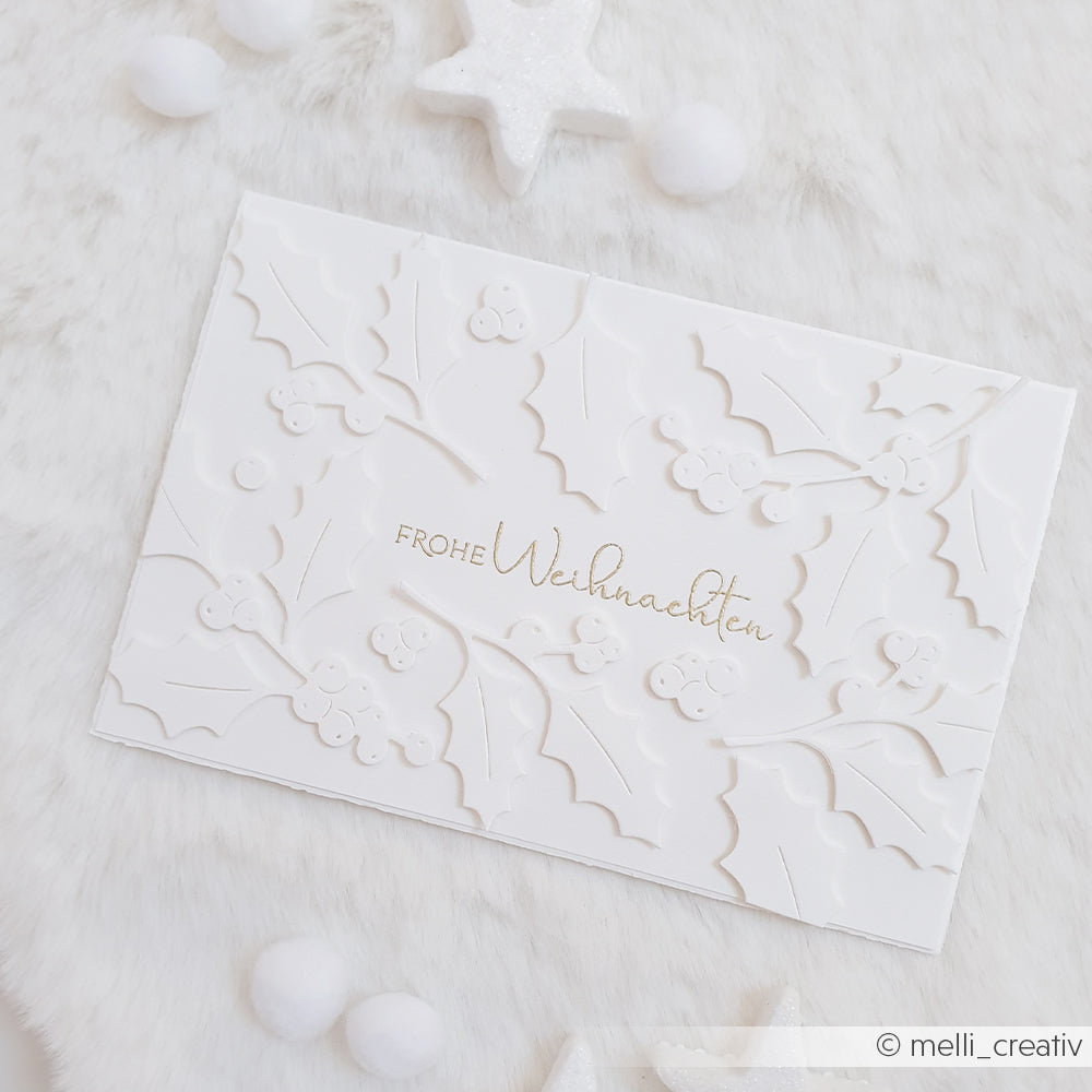 Weiße Karte mit geprägten Stechpalmenblättern (Ilex Zweige) und goldfarbener Aufschrift Frohe Weihnachten.
