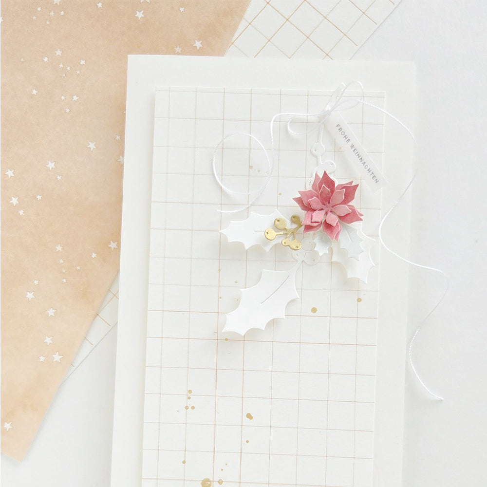Eine Grußkarte mit Karomuster, verziert mit einer Weihnachtsstern-Blüte aus Papier, Stechpalmenzweigen und einem Anhänger mit der Aufschrift „FROHE WEIHNACHTEN“.