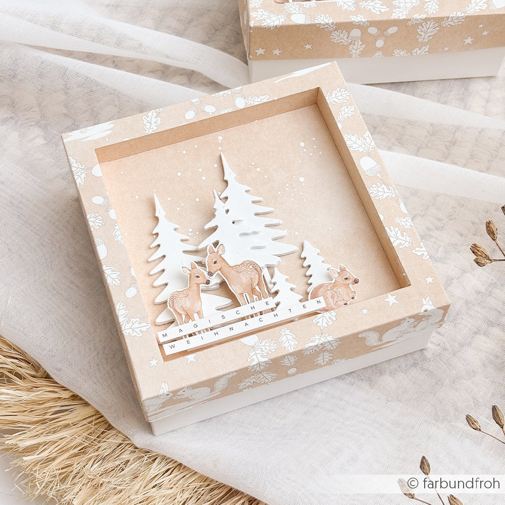 Quadratische Geschenkbox mit Papierausschnitten von Kleine Tannen – Rehe in einem verschneiten Wald –, beschriftet mit Magische Weihnachten.