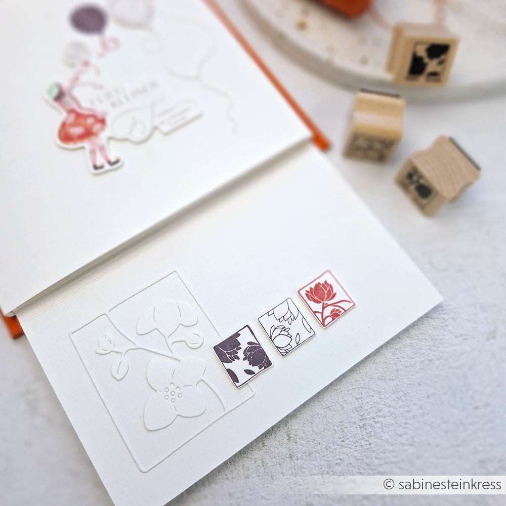 Eine handgefertigte Grußkarte von Alexandra Renke mit Blumenstempeln und Blütenornamentausschnitten, platziert auf einer weißen Fläche mit Holzstempelblöcken in der Nähe.