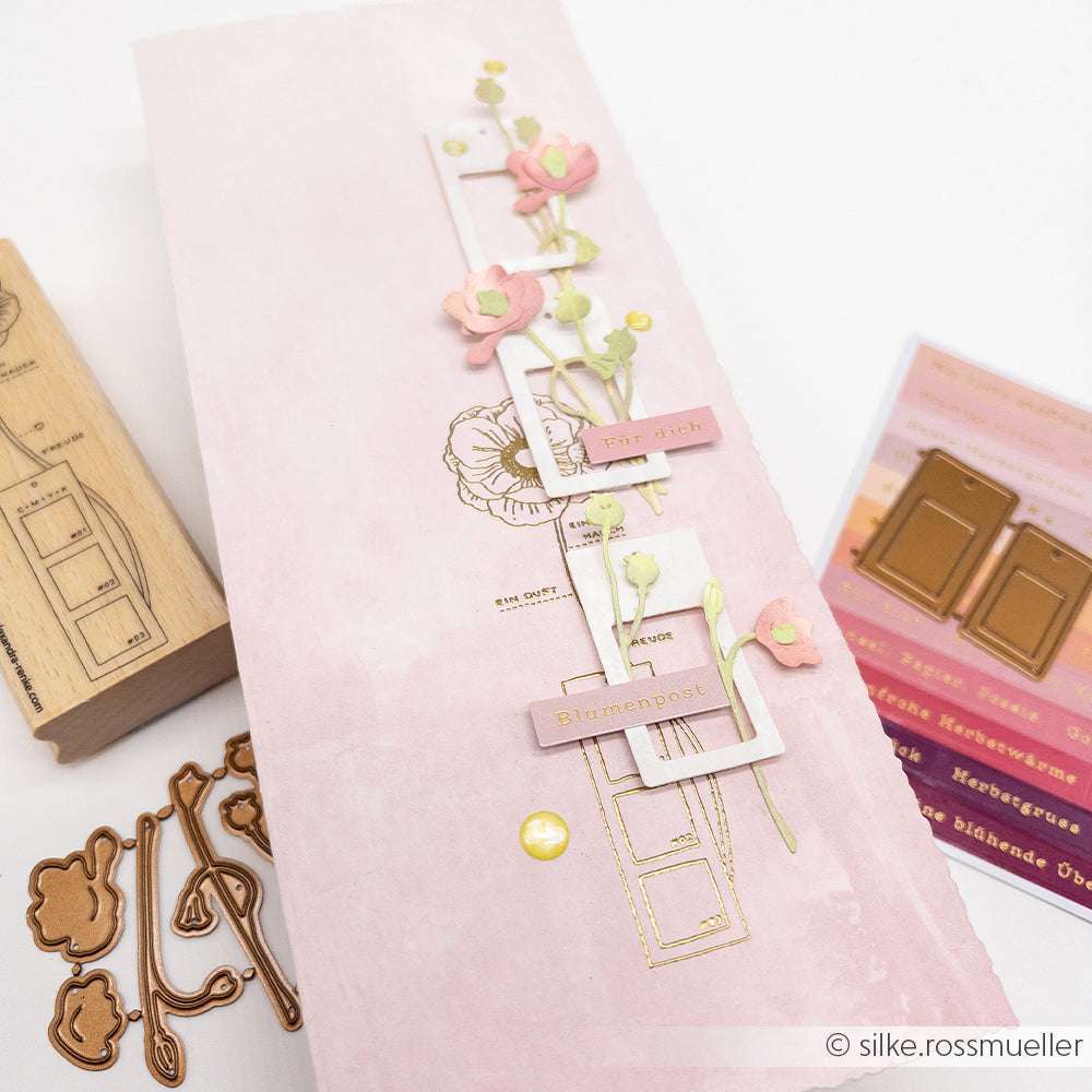 Eine handgefertigte rosafarbene Blumenkarte von Alexandra Renke mit gestanzten Rahmen, Blumen und deutschem Text, der mit Holzstempel und Stempelkissen gestempelt wurde, ausgestellt inmitten von Bastelmaterial.
