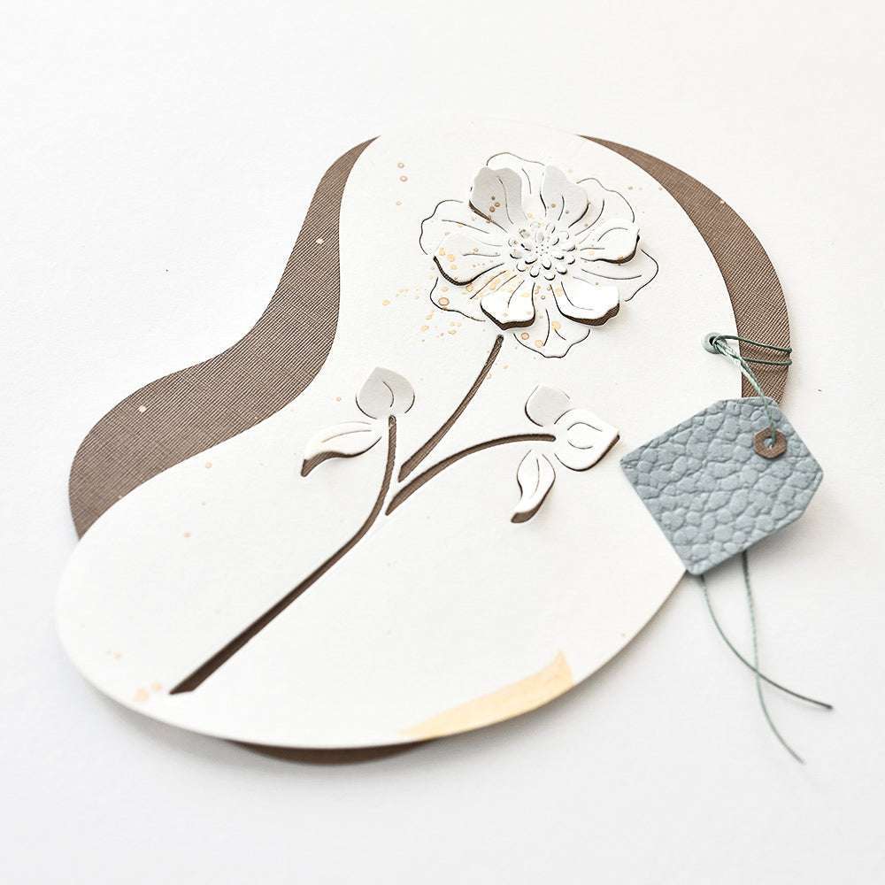Gebogene cremefarbene Karte mit einem ausgeschnittenen Blumenmotiv, das mit der Motivstanze Negativ-Blume 4 von Alexandra Renke hergestellt wurde, sowie einem kleinen grauen, strukturierten Anhänger, der mit Schnur gebunden wird.