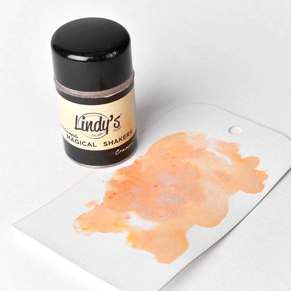Ein kleines Döschen mit Lindy's Stamp Gang Magical Shakers in Crumpet Crumbs, mit einem orangefarbenen Aquarellmuster auf einem weißen Anhänger daneben.