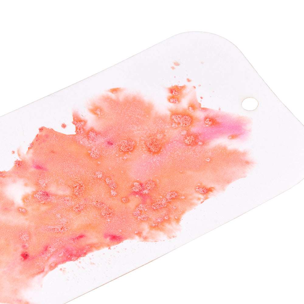 Ein weißer Anhänger mit einem Spritzer rosa und orangefarbenem Lindy's Stamp Gang Magical Powder-hochpigmentiertem Pulver für einen glitzernden Touch und einem kleinen Loch am Rand.