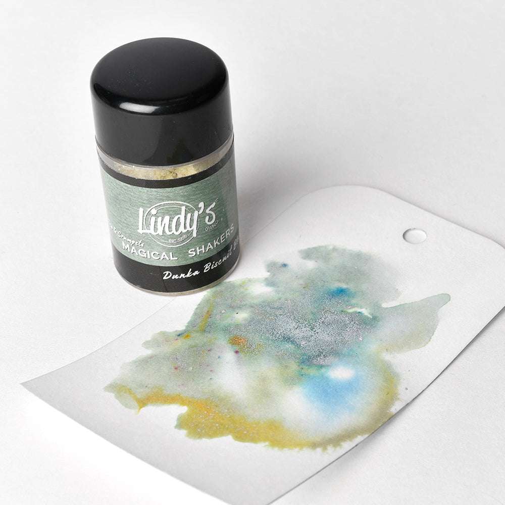 Eine Lindy's Stamp Gang Magical Shaker-Flasche steht neben einem Anhänger mit bunten Aquarellfarben, die leuchtende Farbmischungen hervorheben - ideal zum Testen von Magical Powder-Effekten auf weißem Hintergrund.