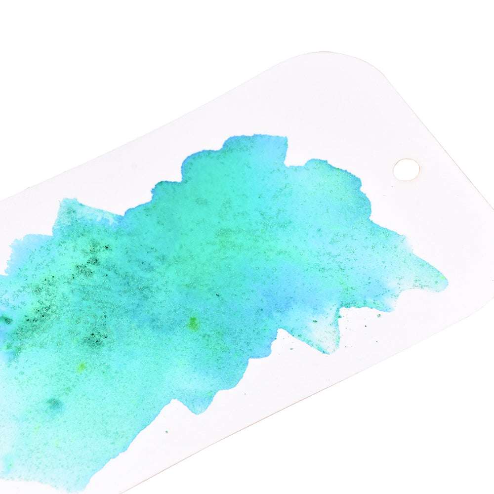 Ein weißer Anhänger mit Loch ist mit einem blau-grünen Aquarellspritzer von Lindy's Stamp Gang versehen, der sich wie von Zauberhand über seine Oberfläche ausbreitet.