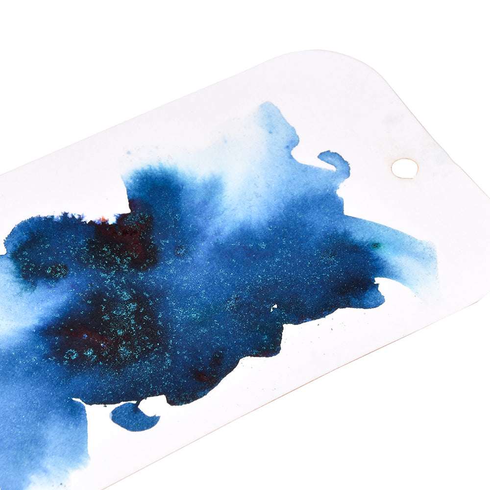 Ein weißes Etikett mit einem Loch und einem großen, abstrakten blauen Tintenklecks - die Pigmente und dunklen Teal-Töne lassen die Farbgebung wie magisches Pulver im Wasser entfalten und erinnern an das kreative Flair von Lindy's Stamp Gang.