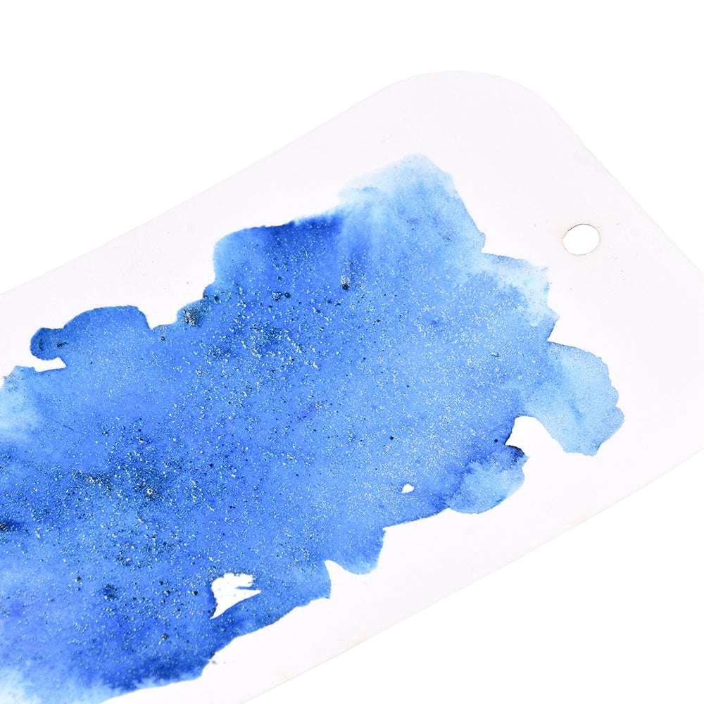 Ein Stück blau schimmernde Aquarellfarbe von Lindy's Stamp Gang, hergestellt mit magischem Pulver, auf einem weißen Papieranhänger mit einem kleinen runden Loch.