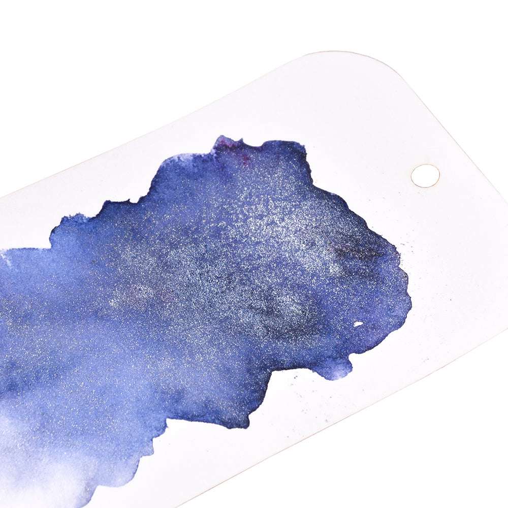Ein unregelmäßiger Fleck aus schimmernder blauer Wasserfarbe, hergestellt mit Lindy's Stamp Gang Magical Powder, ziert einen weißen Anhänger mit einer gestanzten Ecke - ideal für Papierprojekte.