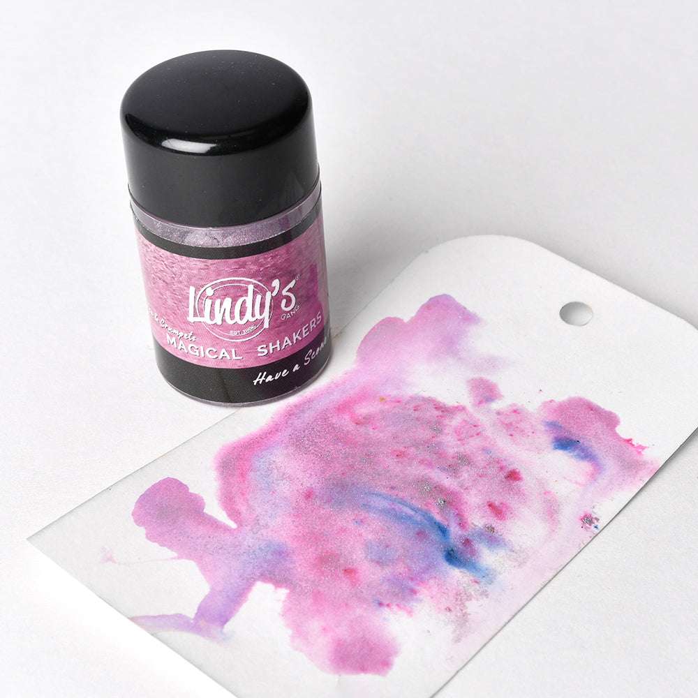 Eine Dose Lindy's Stamp Gang Magical Powder Shakers steht neben einem Schild mit rosa und violetten Mischungen, die mit diesem hochpigmentierten Pulver hergestellt wurden, das sich perfekt für Aquarellmalerei eignet.