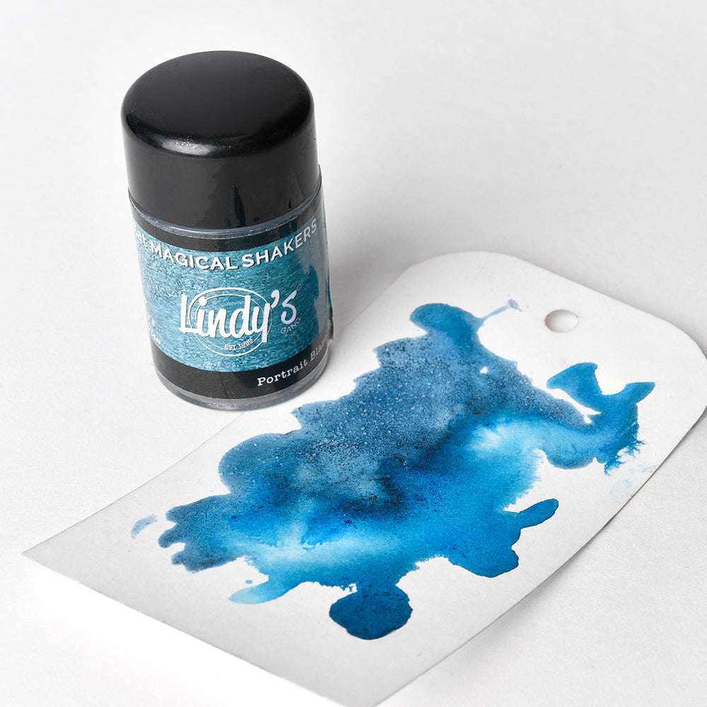 Eine Dose Lindy's Stamp Gang Powder Shaker neben einem Anhänger mit einem blauen Aquarelltintenklecks, der die einzigartige Farbe und hohe Pigmentierung dieses Pulvers hervorhebt.
