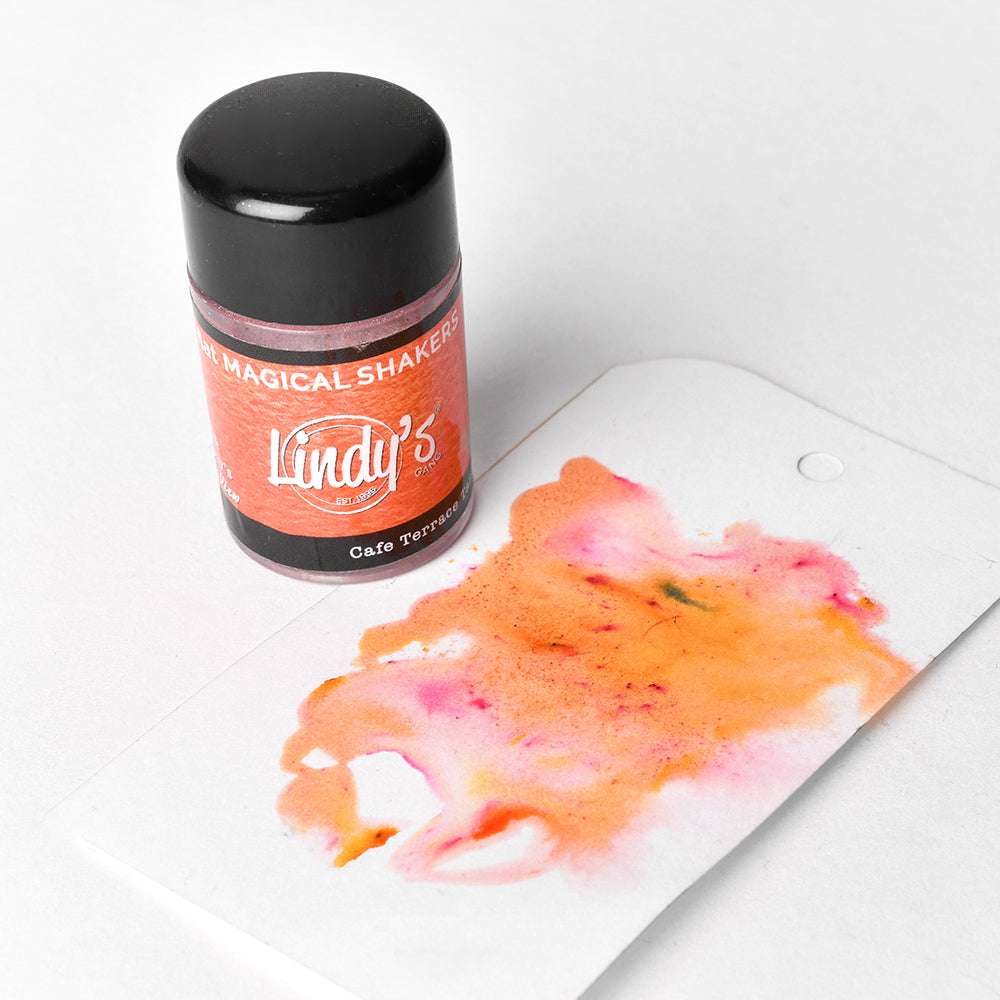 Eine Flasche Lindy's Stamp Gang-Puder steht neben Papier mit orangefarbenen und rosafarbenen Aquarellfarben und zeigt hochpigmentiertes Puder für leuchtende, matte Effekte.