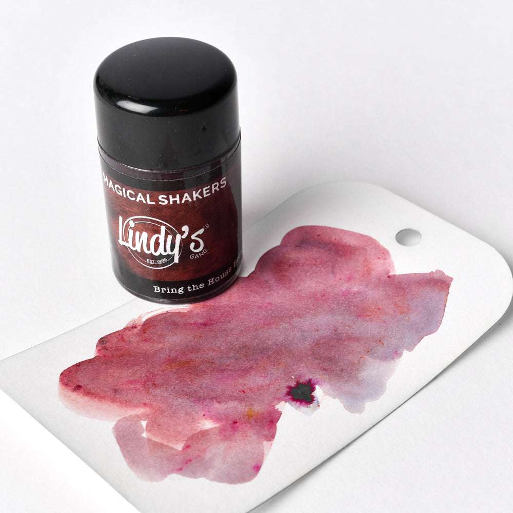 Eine Dose Lindy's Stamp Gang Magical Shakers, gefüllt mit hochpigmentiertem Pulver, steht neben einem weißen Anhänger, auf dem eine rosa-violette Aquarellfarbe abgebildet ist.