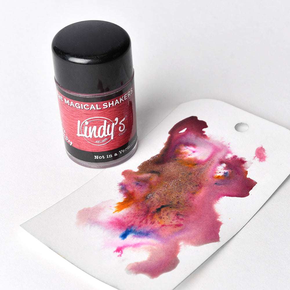 Ein Streuer von Lindy's Stamp Gang Magicals mit hochpigmentiertem Pulver erzeugt einen einzigartigen Farbeffekt, der einen leuchtenden Tintenfleck auf einem weißen Papieranhänger in der Nähe hinterlässt.