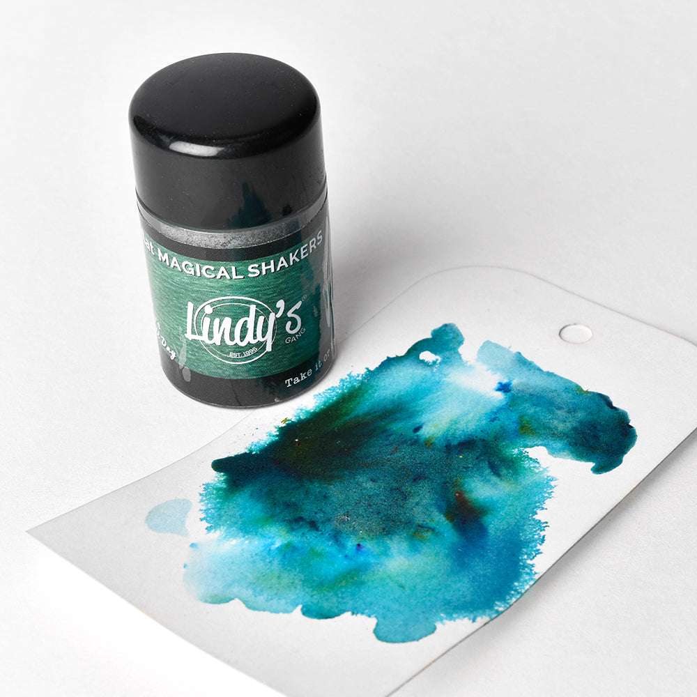 Eine Dose Lindy's Stamp Gang Magical Shakers, gefüllt mit Magical Powder, steht neben einem Anhänger mit blauer und grüner Aquarellfarbe mit Matteffekten.