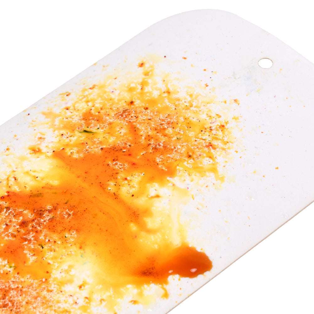 Ein weißes Schneidebrett, das mit orangefarbenen und roten Essensresten, Gewürzen und Spuren des hochpigmentierten Pulvers von Lindy's Stamp Gang befleckt ist.