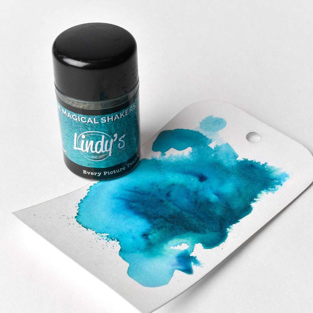 Eine Dose Lindy's Stamp Gang Magical Powder und ein Stück blaue Aquarelltinte auf einem weißen Anhänger, der das hochpigmentierte Pulver für atemberaubende Matteffekte präsentiert.