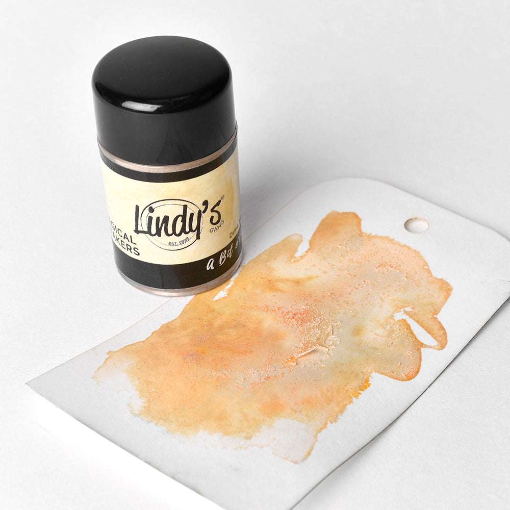 Eine kleine Dose des hochpigmentierten Magical Powder von Lindy's Stamp Gang steht neben einem mit Farbe bespritzten Anhänger mit leuchtenden orangen und gelben Farbmischungen.