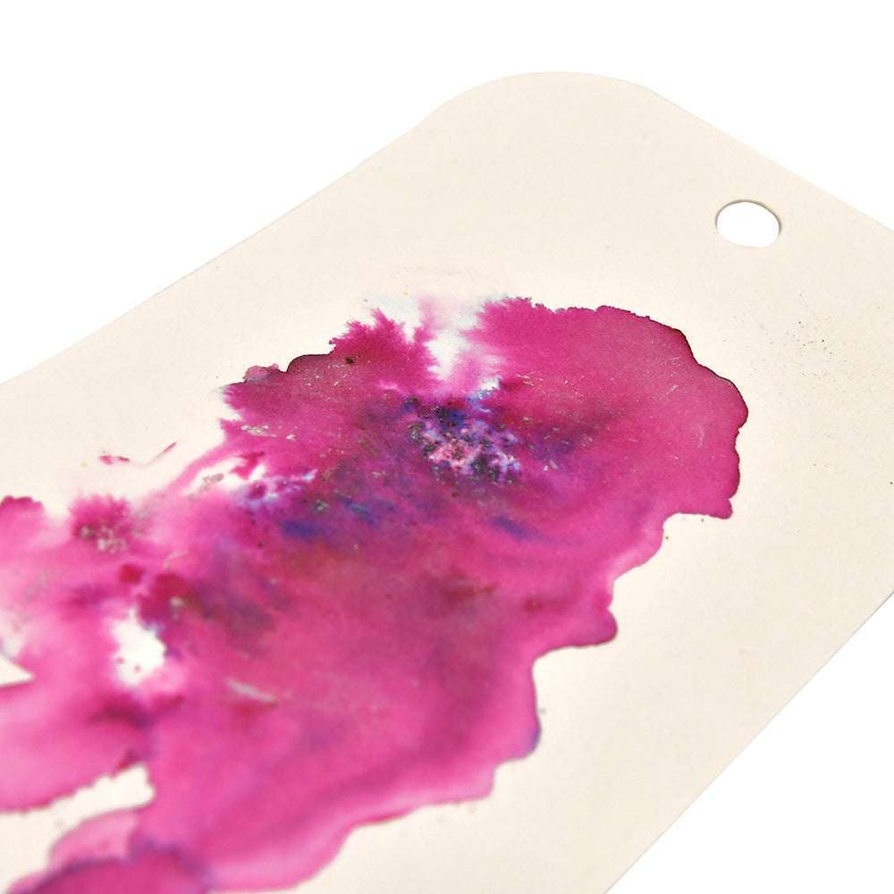 Ein cremefarbener Anhänger mit rosafarbenen und violetten Aquarell-Akzenten, der mit Lindy's Stamp Gang hochpigmentiertem Pulver Magical Powder und einem in eine Ecke gestanzten Loch gestaltet wurde.