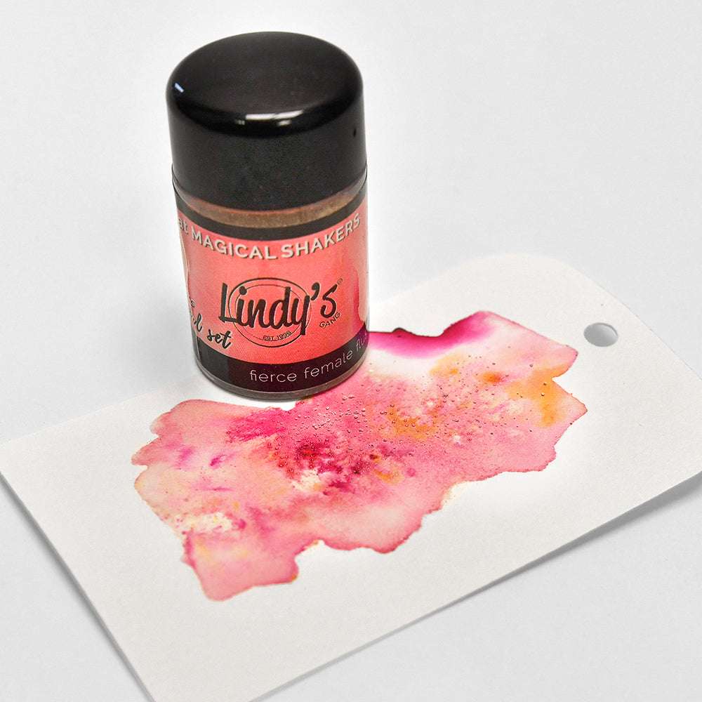 Eine Flasche Lindy's Stamp Gang Magical Shakers, gefüllt mit hochpigmentiertem Pulver, sitzt auf einem mit rosa und goldener Aquarellfarbe verzierten Anhänger.