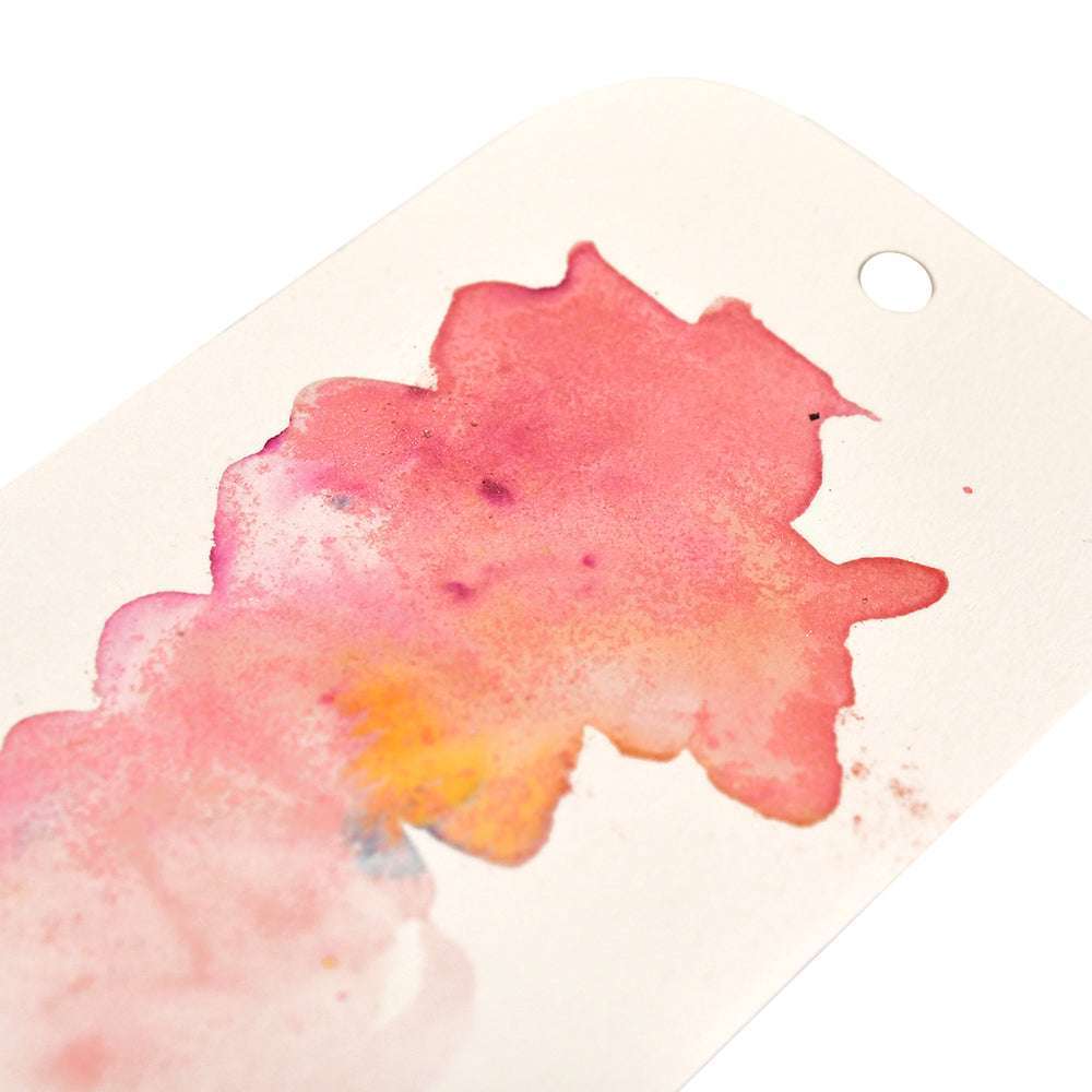 Eine Nahaufnahme eines weißen Anhängers mit einem rosafarbenen und gelben Aquarellfleck - hergestellt mit Lindy's Stamp Gang hochpigmentiertem Pulver Magical Powder - und einem Loch an einem Ende.