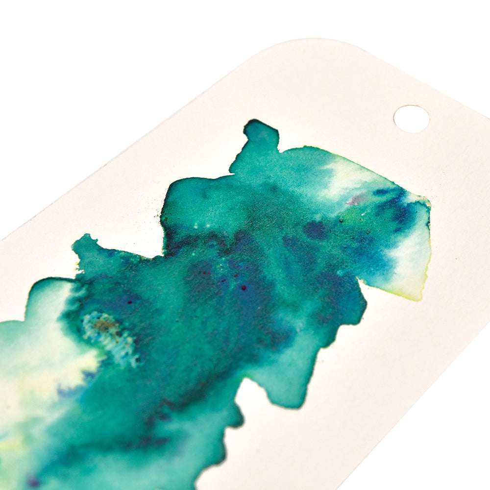 Blaugrüner und grüner Tintenklecks auf einem weißen Papieranhänger mit einem runden Loch oben rechts, der matte Effekte zeigt, die mit den hochpigmentierten Pudern von Lindy's Stamp Gang erzeugt wurden.