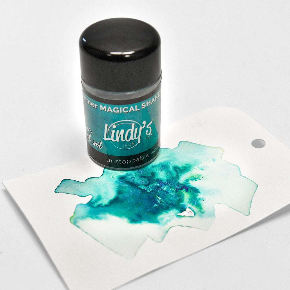 Eine tealfarbene Lindy's Stamp Gang Magical Shaker-Flasche mit leuchtenden Pigmenten ruht auf einer Karte, die einen blau-grünen Aquarellspritzer zeigt, der schöne Farbmischungen hervorhebt.
