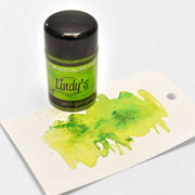 Eine Flasche Lindy's Stamp Gang Magical Powder (hochpigmentiertes Pulver) steht neben einem Anhänger mit einem grünen Aquarellmuster - ideal für kreative Farbmischungen.