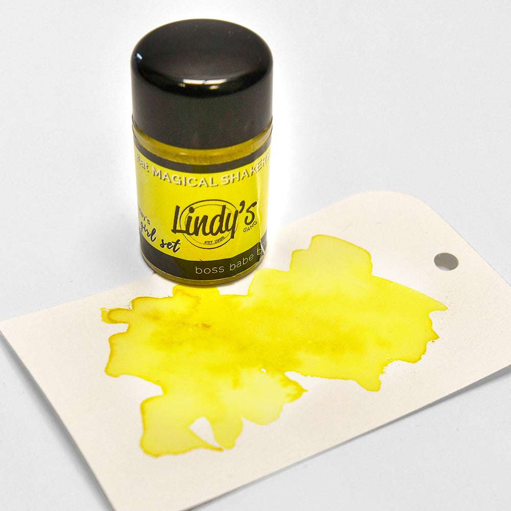Eine gelbe Flasche Lindy's Stamp Gang Magical Shaker mit hochpigmentiertem Pulver und gelber Tinte, die auf ein weißes Schild darunter gespritzt ist.