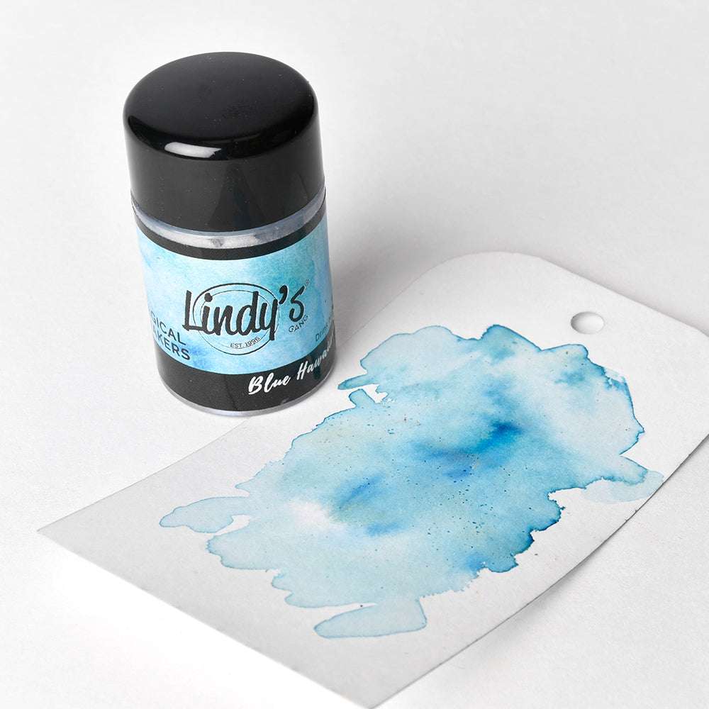 Ein Glas mit Lindy's Stamp Gang Blue Hawaiian Magical Shakers - hochpigmentiertes Pulver - steht neben einem leuchtend blauen Aquarellspritzer auf einem Anhänger.