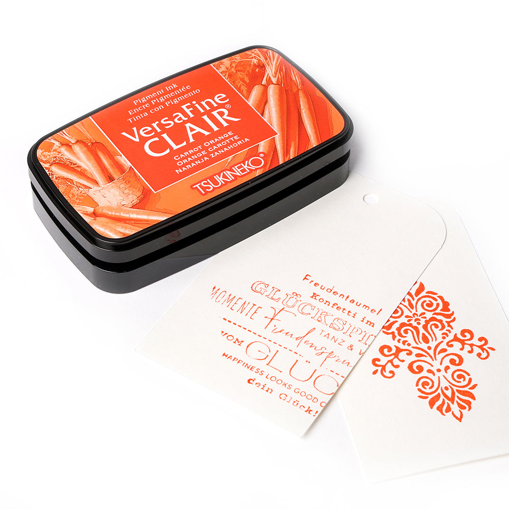 Orangefarbenes Stempelkissen von Tsukineko mit gestempelten Motiven und Text auf zwei weißen Anhängern, die neben dem Stempelkissen angebracht sind.