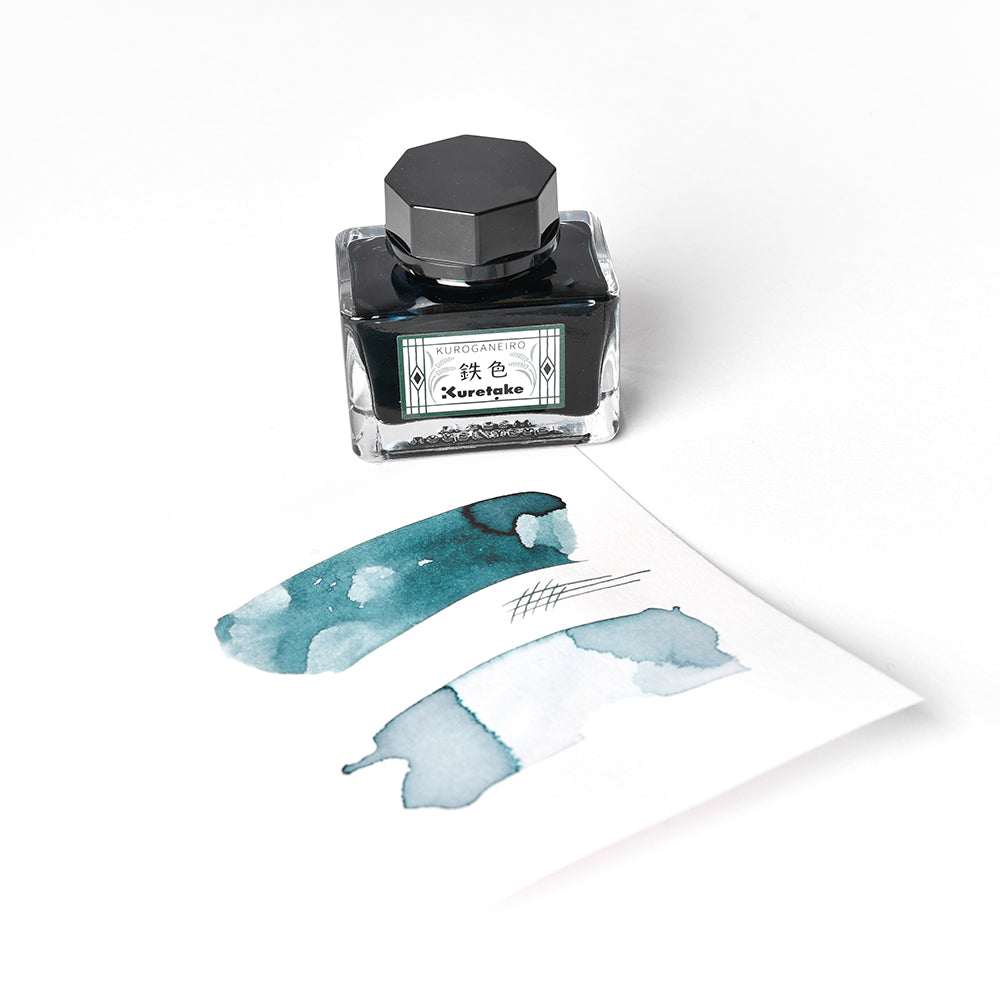 Ein Fläschchen mit Kuretake Kurosame-iro-Tinte aus der Serie MEIJI NO IRO steht neben einem Blatt mit blauen Tintenklecksen und Kritzeleien.
