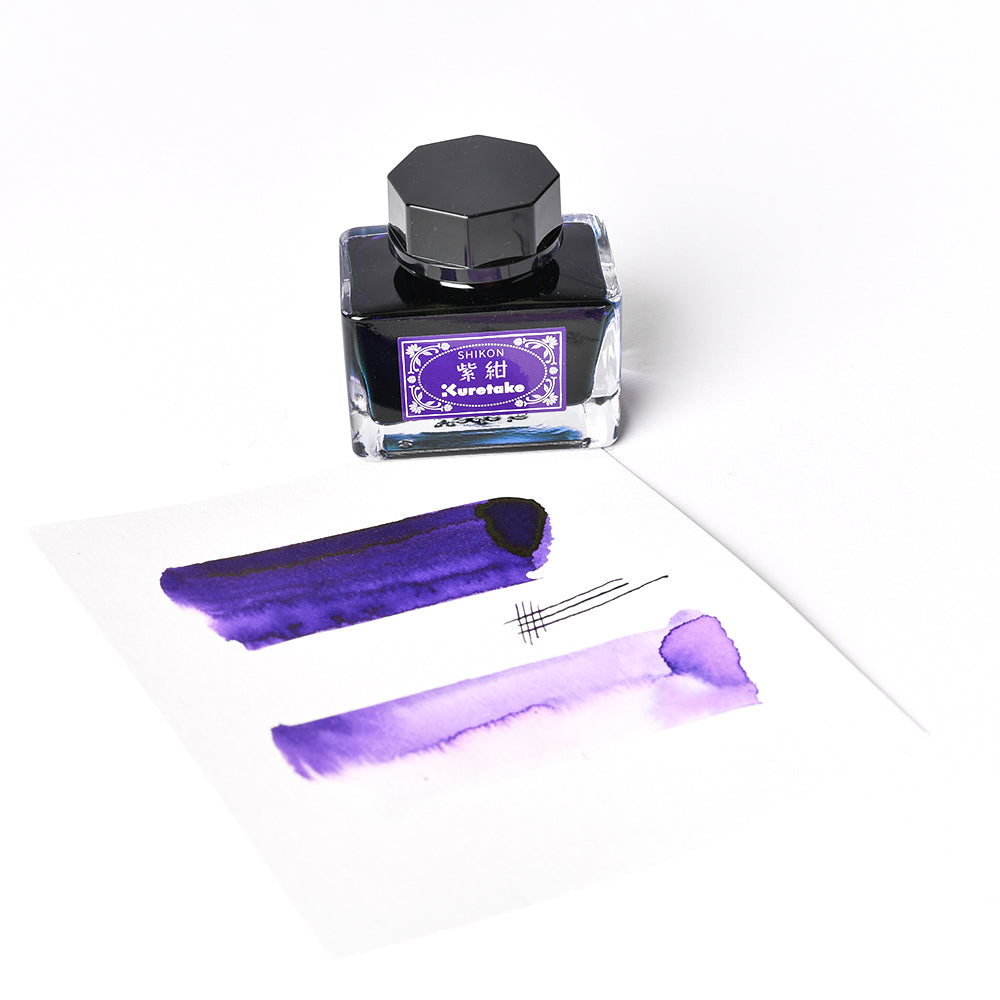 Ein Fläschchen mit Kuretake-Tinte steht neben einem Papier mit leuchtend violetten Flecken und Stiftmarkierungen, die sich alle vor einem klaren weißen Hintergrund befinden.