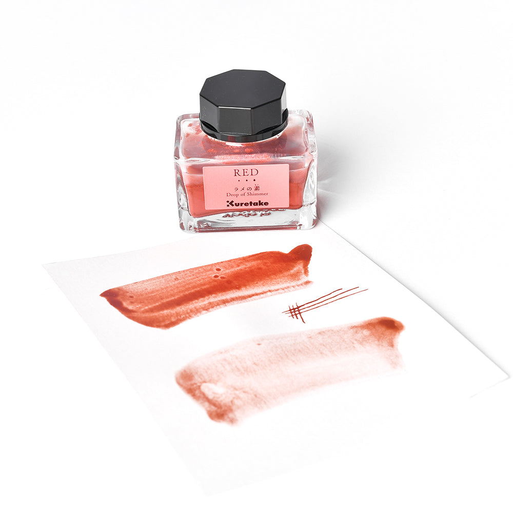 Ein Fläschchen mit roter Kuretake-Tinte und ein Blatt Papier mit roten Farbtupfern und schimmernden Glanzlichtern stehen auf einem weißen Hintergrund.