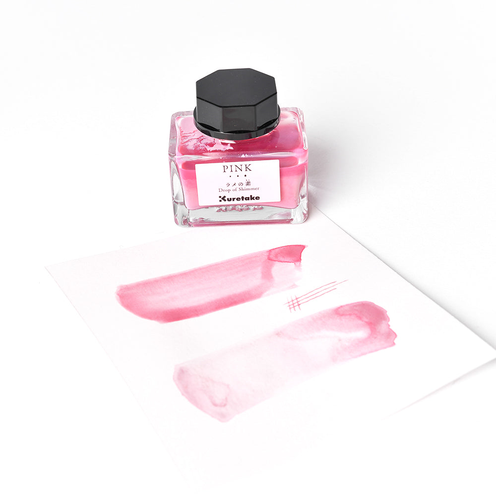 Ein Fläschchen mit rosafarbener Tinte von Kuretake, ideal für Tinte mischen, steht neben weißem Papier mit rosafarbenen Mustern und schimmernden Linien von Schimmertinte.
