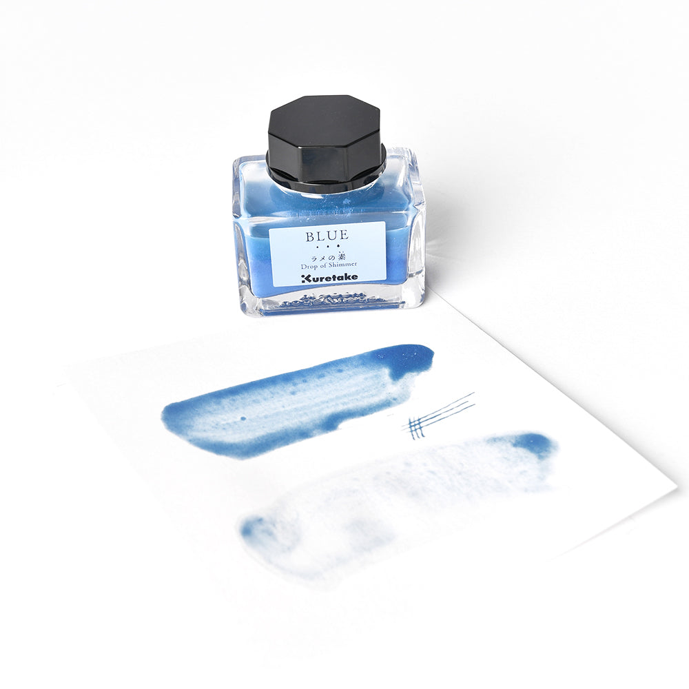 Ein Fläschchen mit blauer Kuretake-Tinte mit schimmernden Akzenten, die auf einem weißen Blatt Papier aufgetragene Flecken und funkelnde Linien mit dezentem perlenartigem Glitzer zeigt.