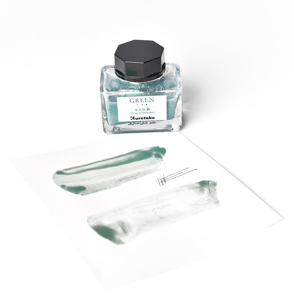 Ein Fläschchen mit grüner Kuretake-Tinte und Tinte Schimmer steht neben einem Blatt Papier, das mit grünen Tintenklecksen und Stiftspuren bedeckt ist, alles auf weißem Hintergrund.