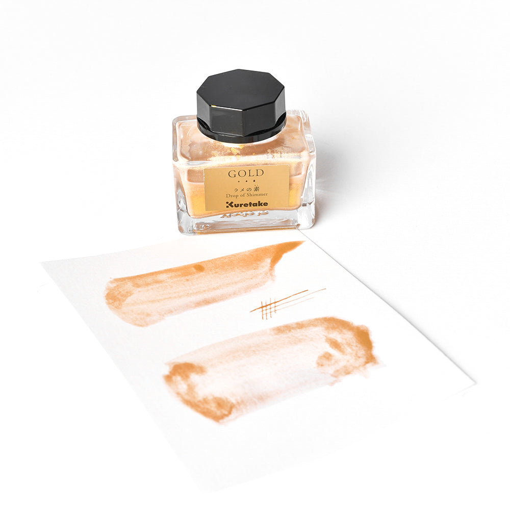 Ein Glasfläschchen mit goldener Kuretake-Tinte, die für ihren schimmernden Effekt bekannt ist, steht neben einem Papier, das mit perlmuttfarbenen Glitzerflecken und leuchtenden Linien auf weißem Hintergrund verziert ist.