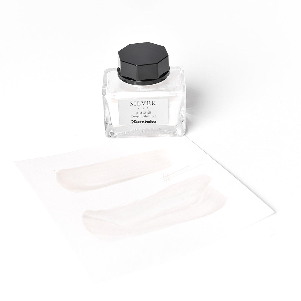 Ein Fläschchen mit silbern schimmernder Kuretake-Tinte mit schwarzer Kappe steht neben einem Papier, das zwei perlenartige Pinselstriche zeigt.