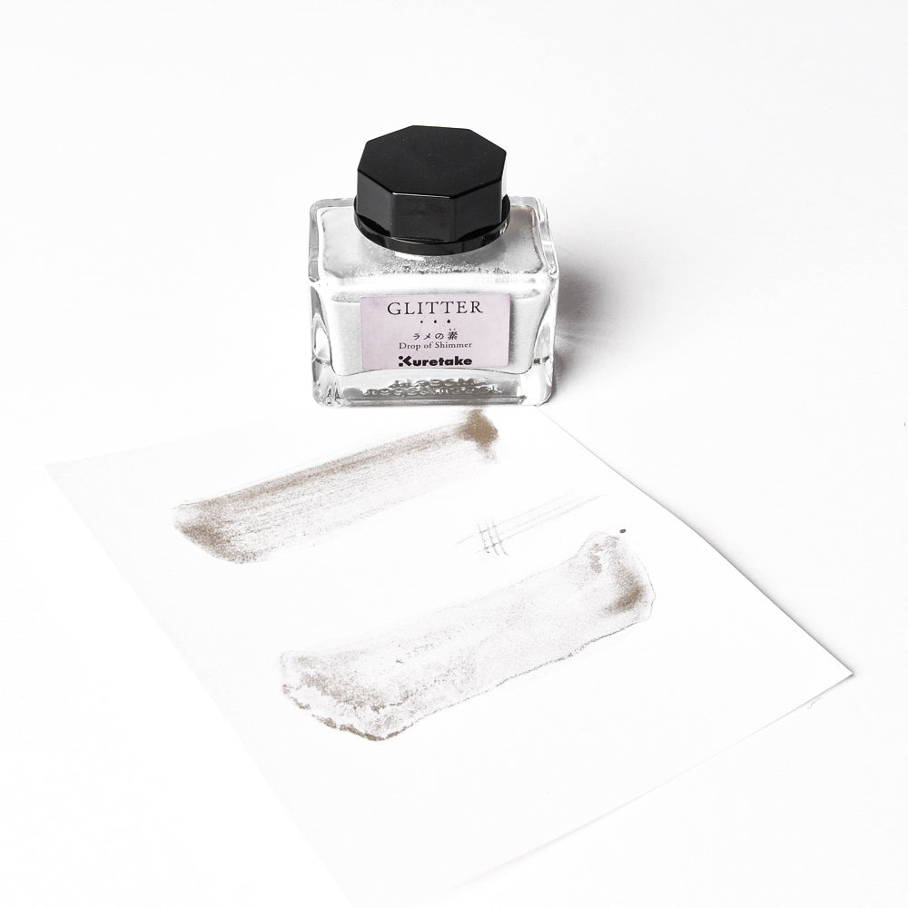 Ein Fläschchen mit silberner Glitzertinte von Kuretake, die für ihren perlmuttartigen Glitzer bekannt ist, steht neben einem Blatt mit schimmernden Silberflecken auf weißem Hintergrund.