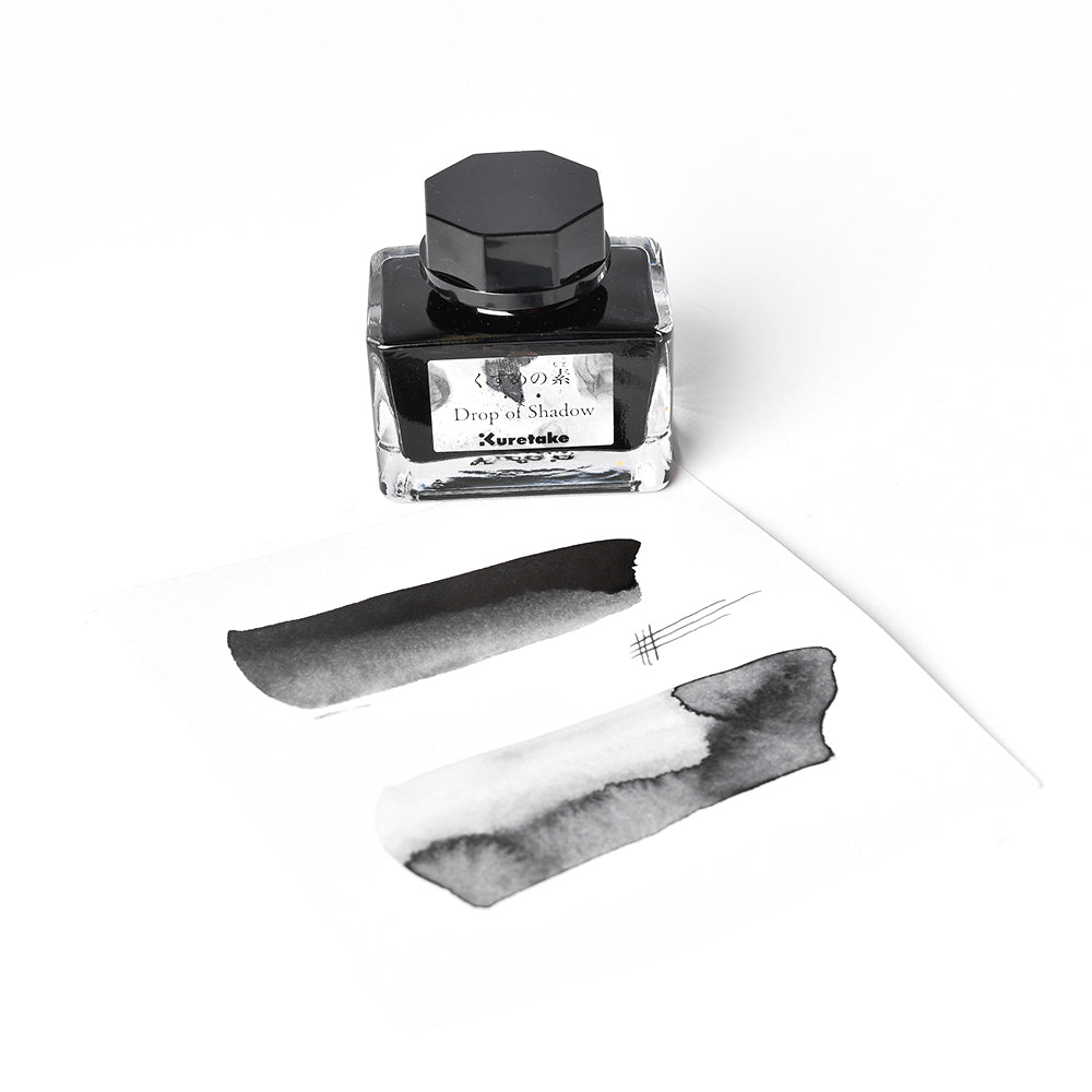 Ein Fläschchen mit Kuretake-Tinte steht neben weißem Papier und zeigt zwei schwarze Flecken, drei feine Linien und einen subtilen Schattenwurf-Effekt, der verdeutlicht, wie dunkle Farben mischen eine beeindruckende Tiefe erzeugen.