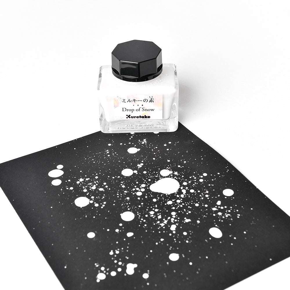 Ein Fläschchen mit weißer Tinte von Kuretake Drop of Snow und der Aufschrift "Deckkraft Tinte erhöhen" steht neben Farbspritzern auf schwarzem Papier, die mit ihrer matten Oberfläche einen auffälligen Kontrast bilden.