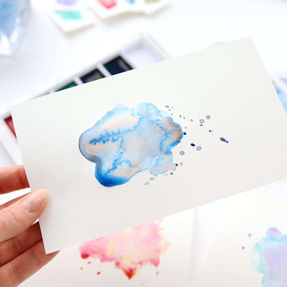 Eine Hand hält eine Karte mit einem blauen Aquarellfarbenspritzer; im Hintergrund ist eine Kuretake Gansai Tambi Farbpalette zu sehen.