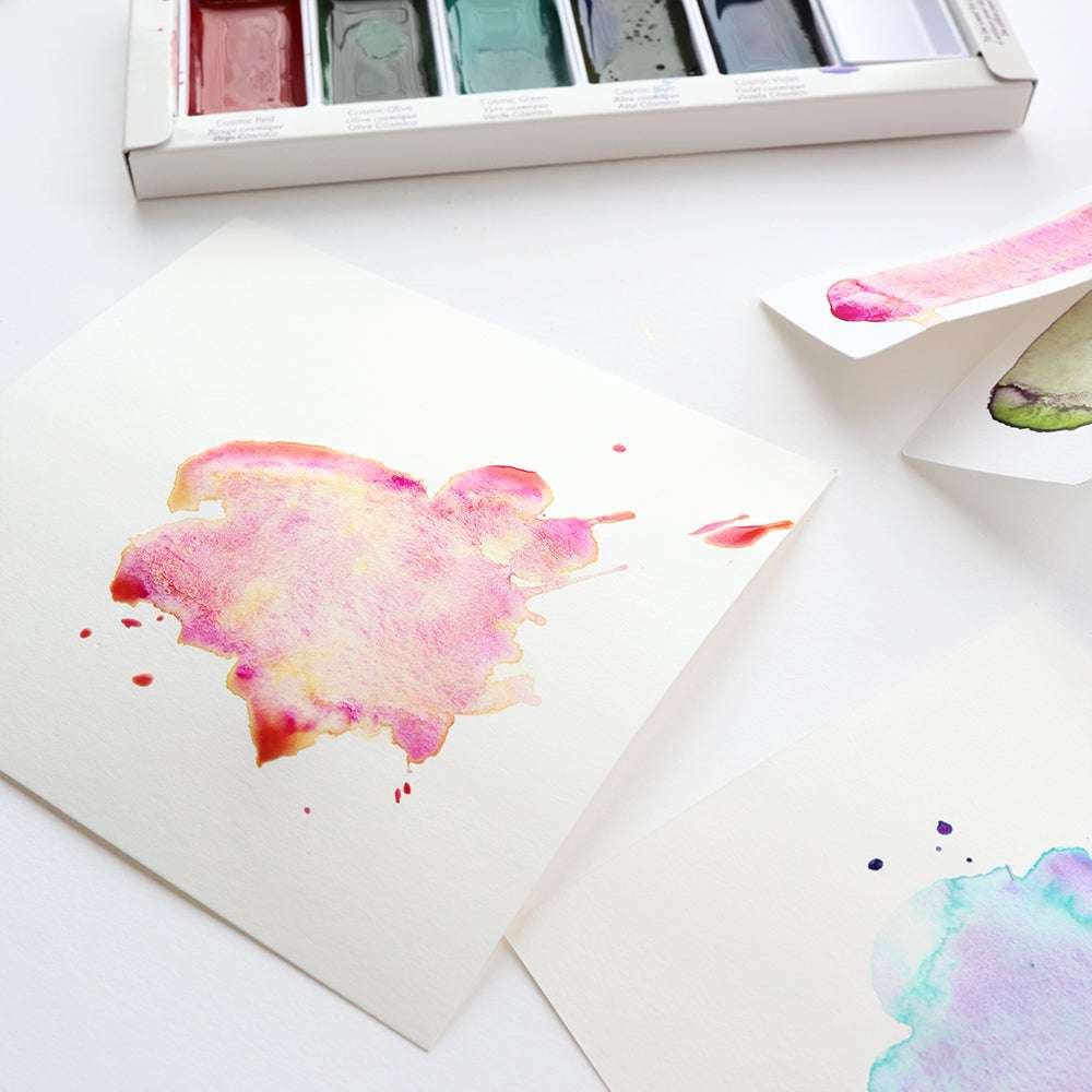 Ein rosafarbenes Aquarell auf Papier ist neben einem geöffneten Set von Kuretake Gansai Tambi-Farben und anderen bemalten Blättern ausgestellt.