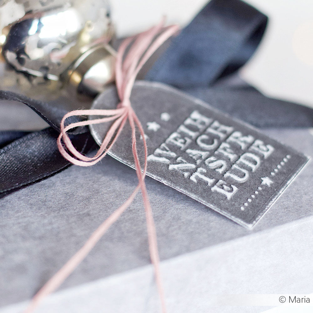 Nahaufnahme eines Geschenks mit silbernem WEIHNACHTSFREUDE-Anhänger, gebunden mit pinkfarbenem ITO Garn für kreative Projekte und einem schwarzen Band - perfekt für eine stilvolle Geschenkverpackung.