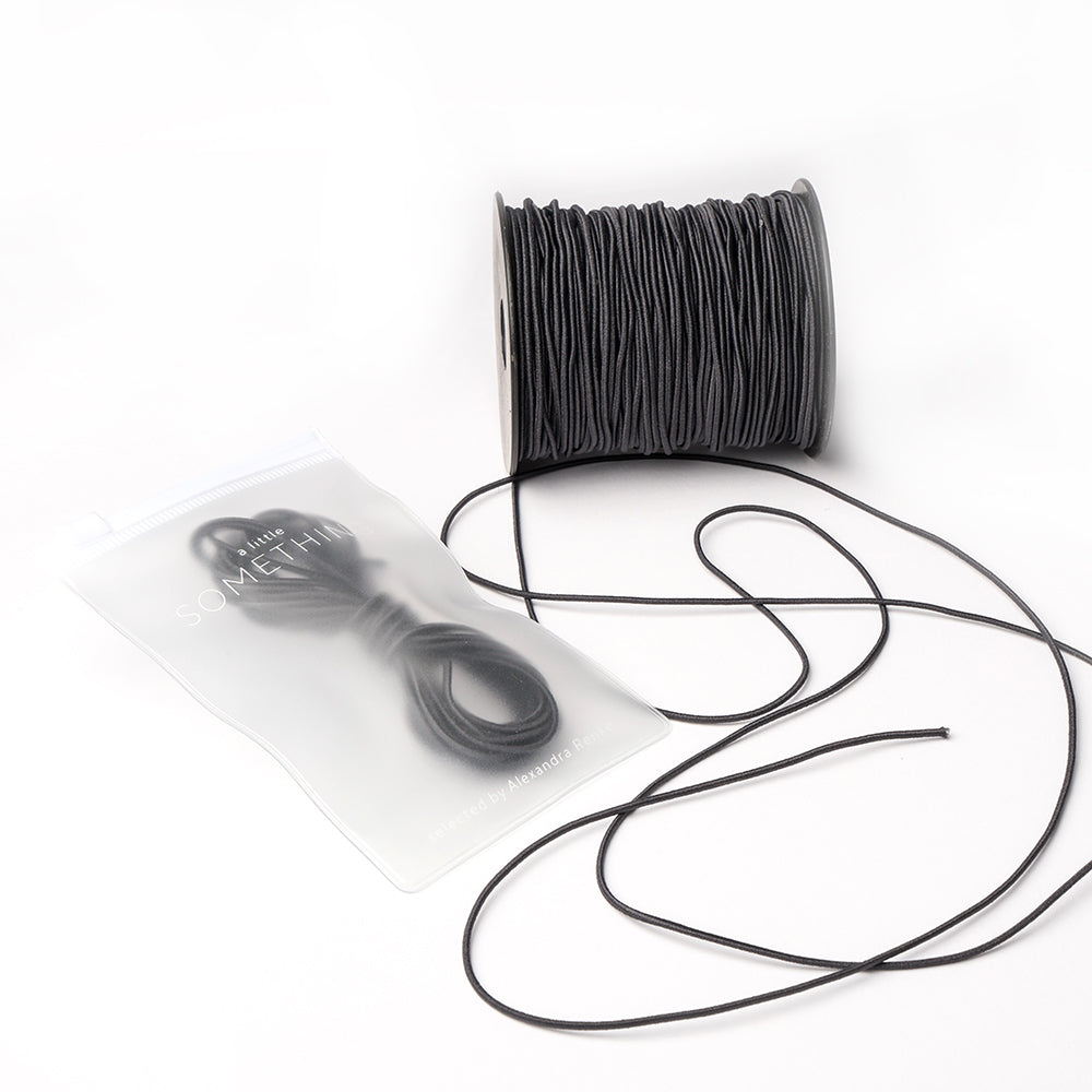 Eine Spule schwarzes Elastikband von Alexandra Renke liegt neben einem wiederverschließbaren Beutel mit zusätzlicher Kordel, ideal für das Bastelprojekt, alles auf weißem Hintergrund.