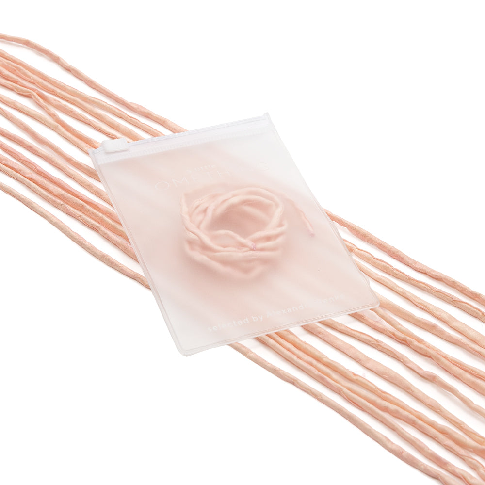 Mehrere lange, blassrosa Garnstränge und ein rundes Stück Garn liegen auf einem durchscheinenden weißen Beutel von ☆, der an handgenähte Geschenkverpackungen erinnert und zart mit einer Seidenschnur verziert ist.